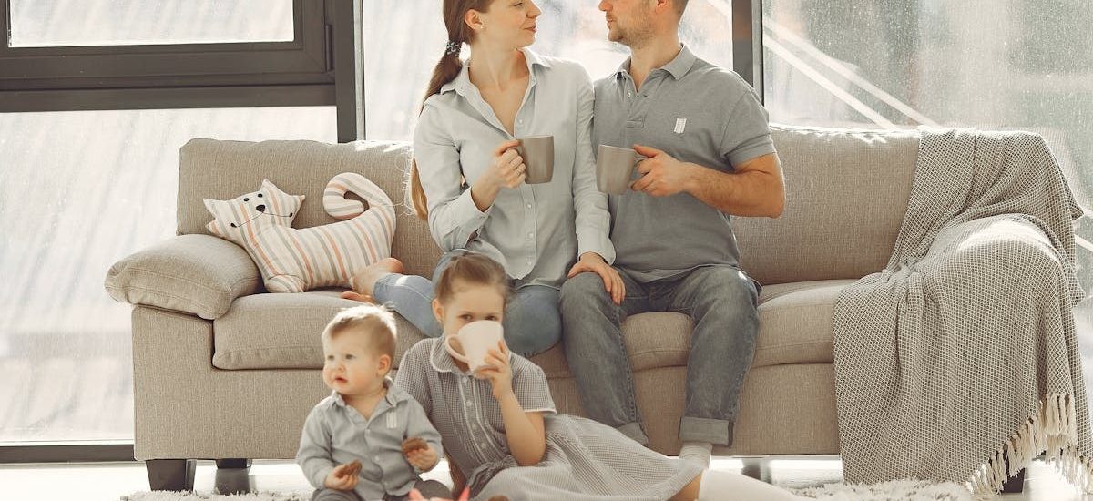 Happy family with children spending time at home while parents sitting on couch with cups of coffee and looking at each other when kids resting on floor