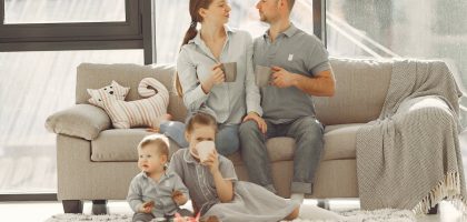 Happy family with children spending time at home while parents sitting on couch with cups of coffee and looking at each other when kids resting on floor
