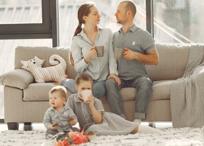 Happy family with children spending time at home while parents sitting on couch with cups of coffee and looking at each other when kids resting on floor